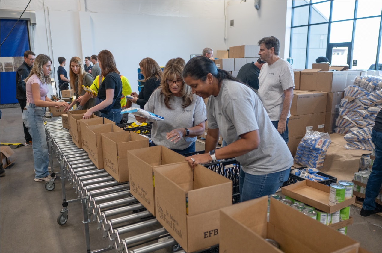 A group of volunteers work together in a warehouse, packing food and supplies into boxes on a conveyor system for community assistance and donation efforts.