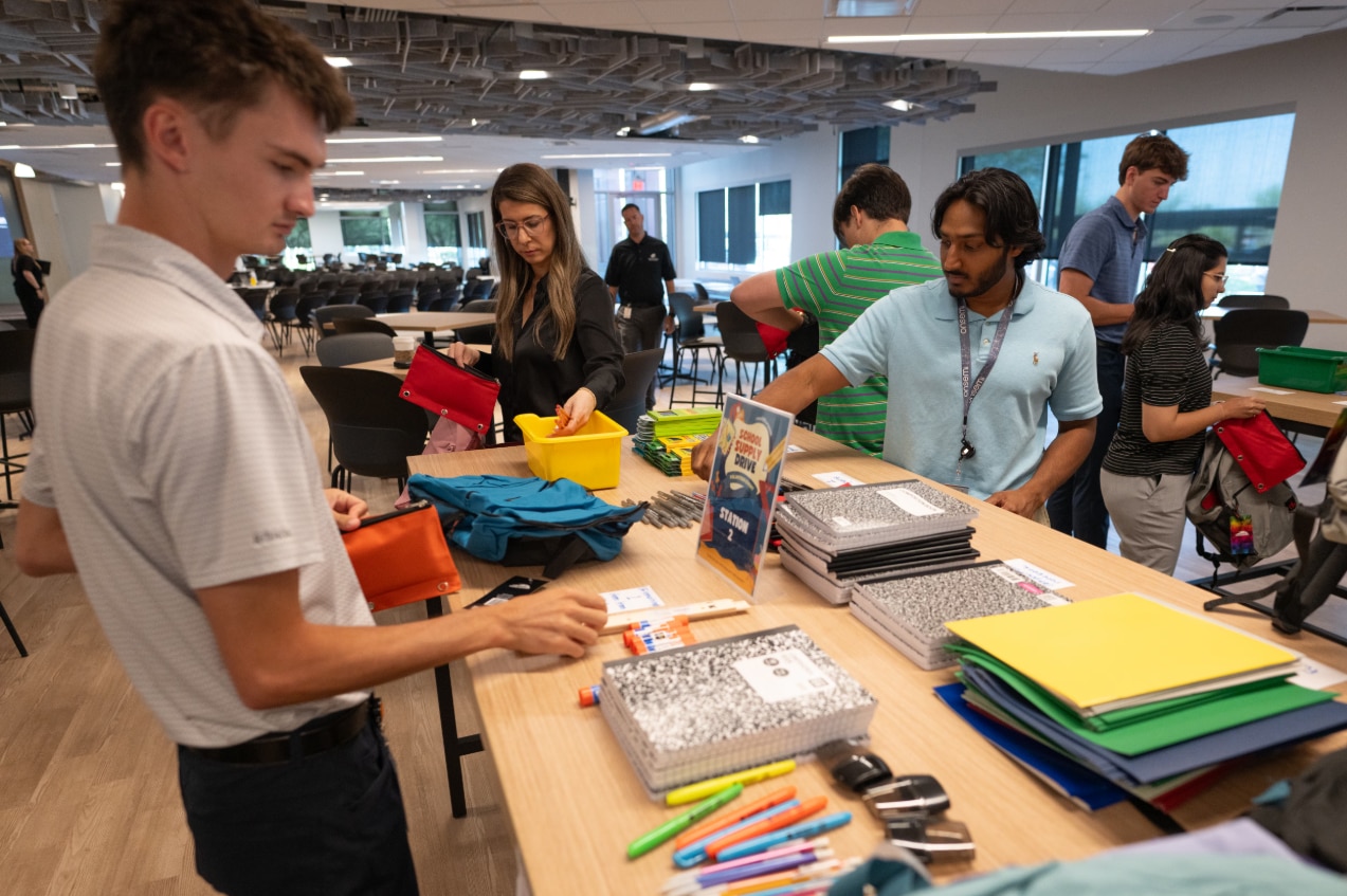 Volunteers sort notebooks, folders, pens, and backpacks at donation stations during a school supply drive in a corporate office setting.