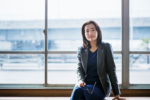 A formally dressed woman sitting by a window, looking into the camera.